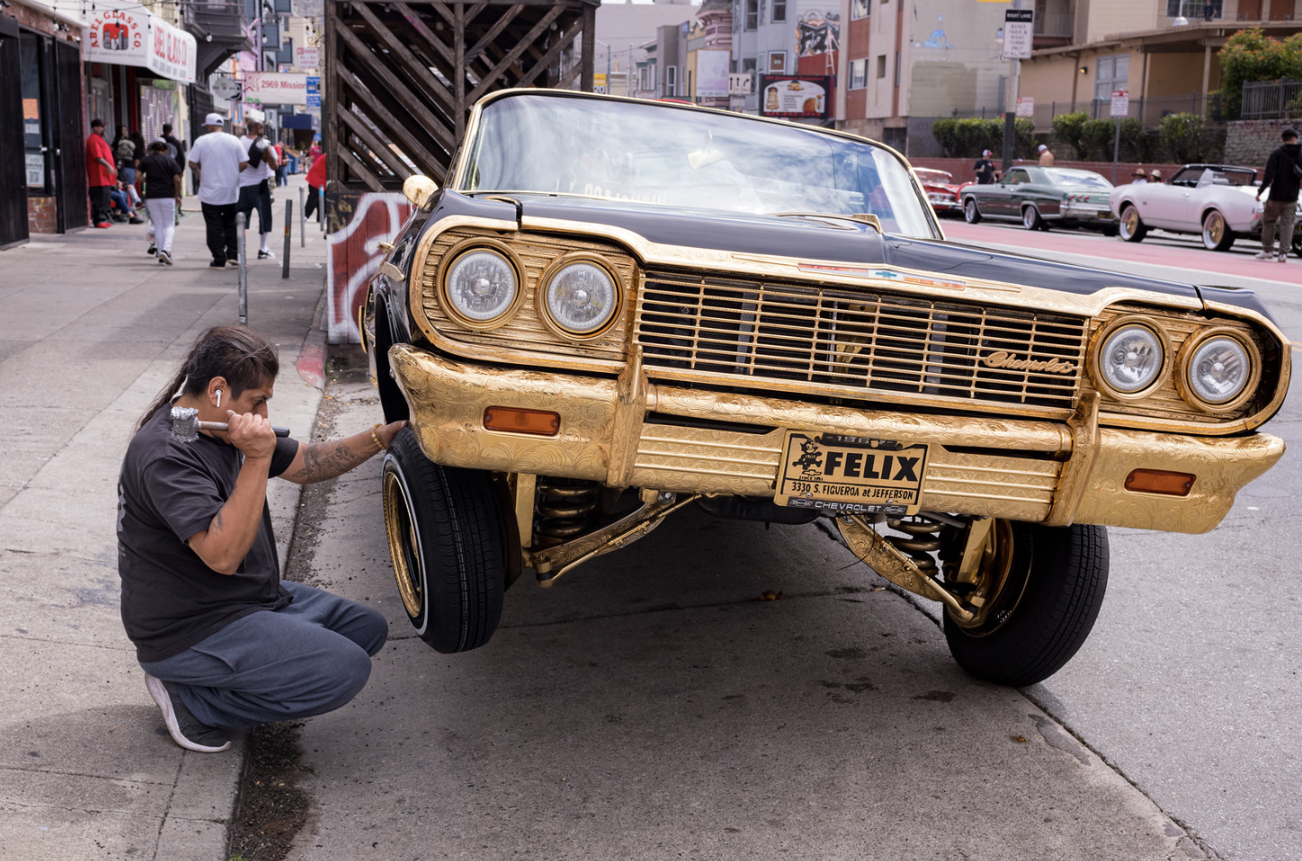 Man squats next to a gold-trimmed car with raised hydraulics on a city street while talking on a phone.